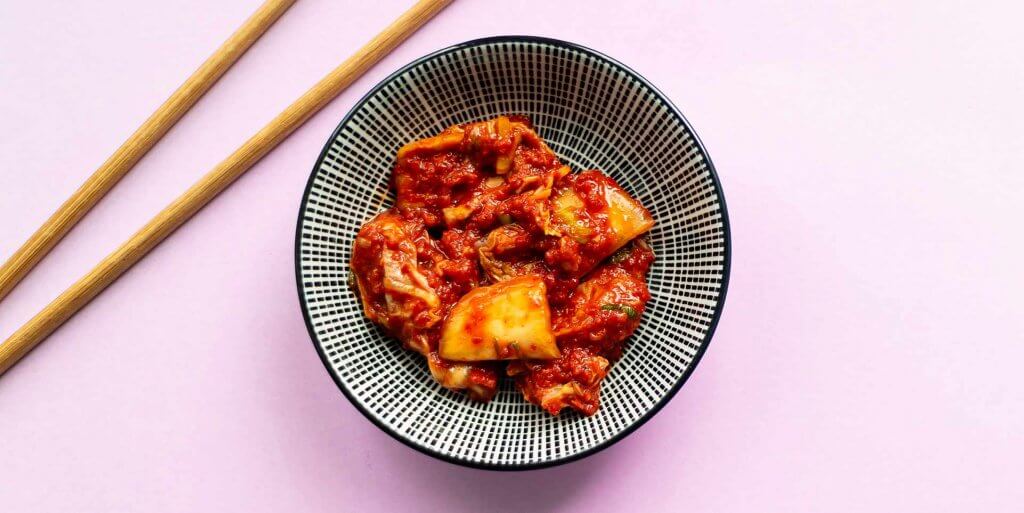 Cabbage kimchi in a small black and white patterned bowl with wooden chopsticks laying to the side